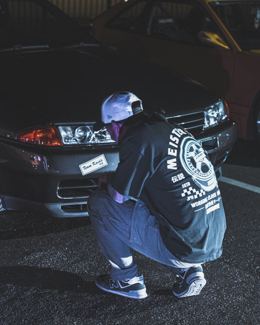 Clubhouse Meister Wheels Tee worn beside a Japanese car at night